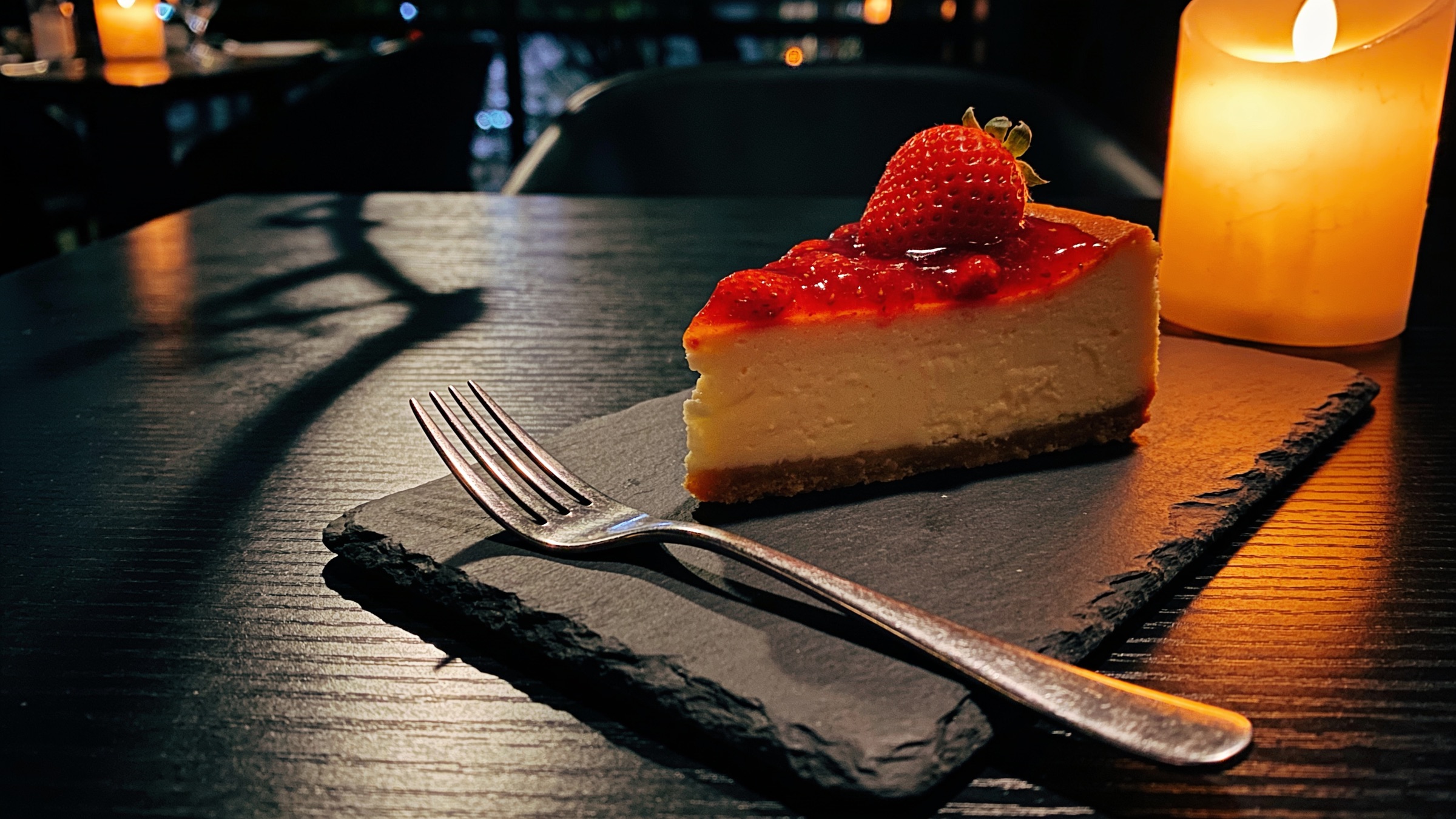 A slice of cheesecake with strawberry topping on a candlelit restaurant table.